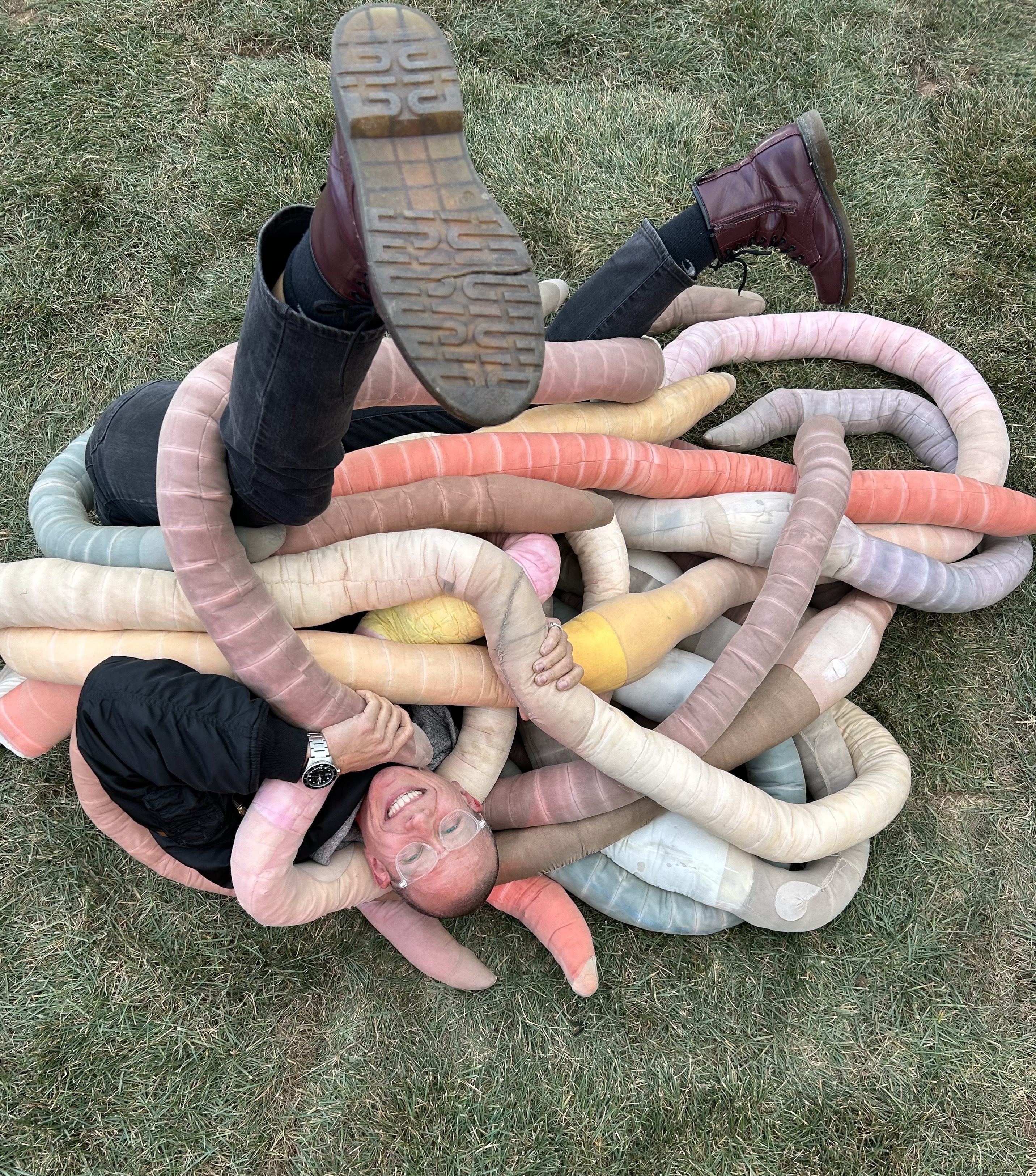 Val is laying on the grass entwined in a pile of large, stuffed, fibre-art worms. He is smiling joyfully at his good fortune.