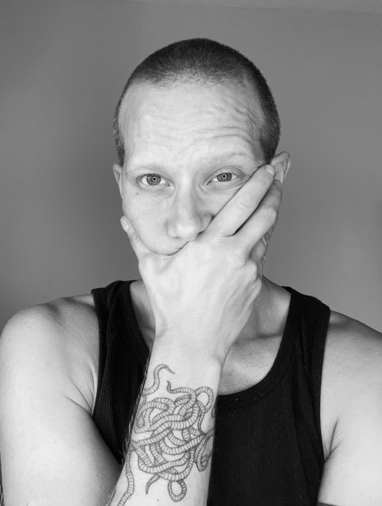 black and white portrait of Val. He is a white man with a shaved head wearing a black tank top. He is covering his mouth with his hand, revealing a wrist-wrapping tattoo of a clew of worms.